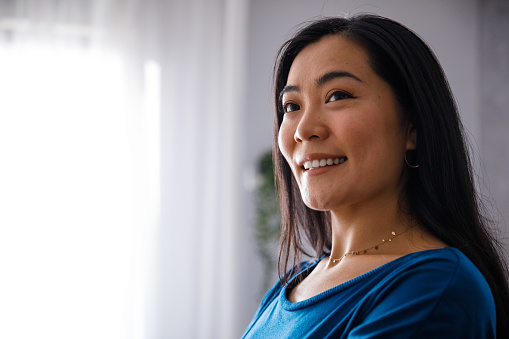 Portrait of beautiful young Mongolian woman standing in her living room, looking away, smiling and contemplating.