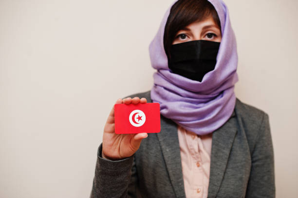 Coronavirus country concept. Portrait of young muslim woman wearing formal wear, protect face mask and hijab head scarf, hold Tunisia flag card against isolated background. Coronavirus country concept. tunisia woman stock pictures, royalty-free photos & images