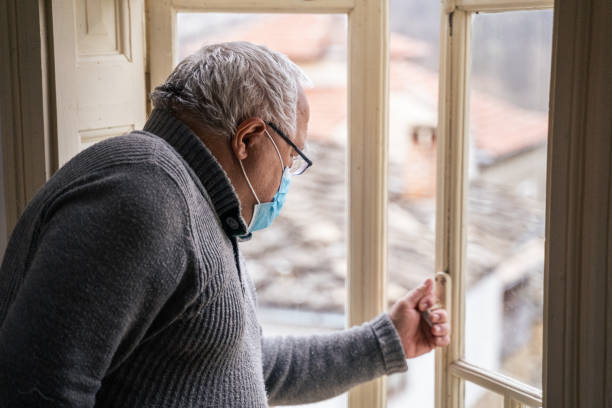 Coronavirus protection. Active senior man looking through window. Wearing mask to avoid infectious diseases while at his home. Portrait of a senior man with protective mask on his face while in outbreak. restraining stock pictures, royalty-free photos & images