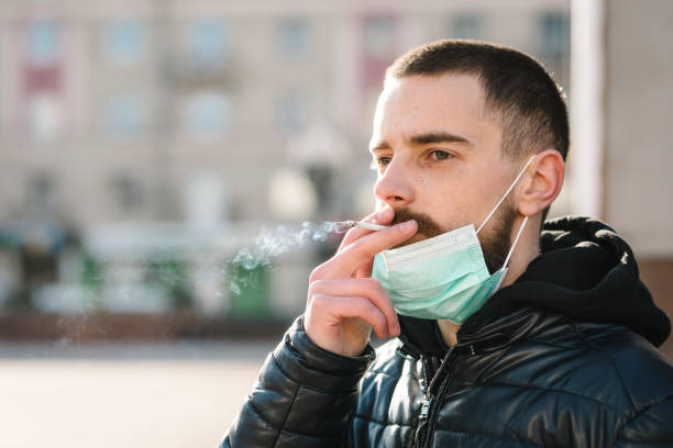Coronavirus. Smoking. Closeup man with mask during COVID-19 pandemic smoking a cigarette at the street. Smoking causes lung cancer and other diseases. The dangers and harm of smoking. stock photo