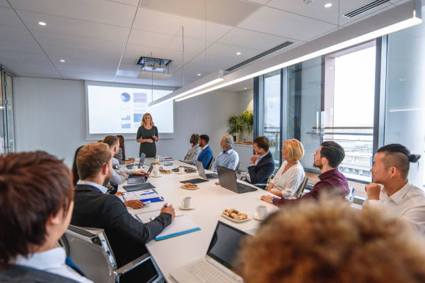 Corporate Executive Team Listening to CEO Presentation Over the shoulder view of executive team listening to mature female CEO presenting project plans for the coming year. board room stock pictures, royalty-free photos & images