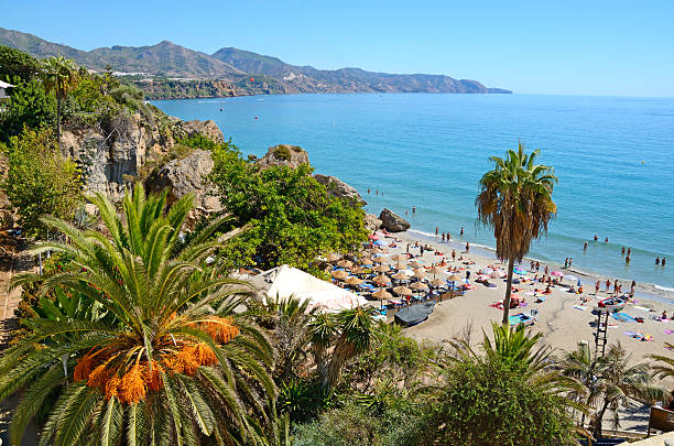 Costa del Sol Nerja, Spain - September 18th, 2016: Coastline in Nerja, famous resort in Costa del Sol, called Balcon de Europe. Mediterranean sea on the right. Sierra Nevada mountains on the horizon. In the foreground on right a little beach with straw sunshades and many people. In the foreground on the left rocks ovegrown with many bush and a date palm. Horizontal image in a sunny day. costa del sol málaga province stock pictures, royalty-free photos & images