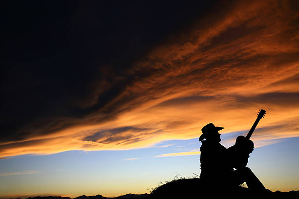 Country Singer Songwriter Silhouette A man playing guitar at sunset. Silhouette. Unrecognizable male singer songwriter sitting on a hay bale outside against beautiful sunset sky. Themes include country music, roots music, festival, artist, singer, singing, songwriter, poet, cowboy, western, bluegrass, Americana, instrument, picker, playing, musician, and alone.  country and western music stock pictures, royalty-free photos & images