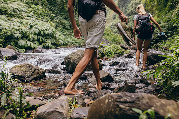 Couple crossing the stream barefooted Low section shot of man and woman crossing the stream barefooted. Couple walking by the creek in forest with their shoes in hand. Hikers hiking in forest. extreme terrain stock pictures, royalty-free photos & images