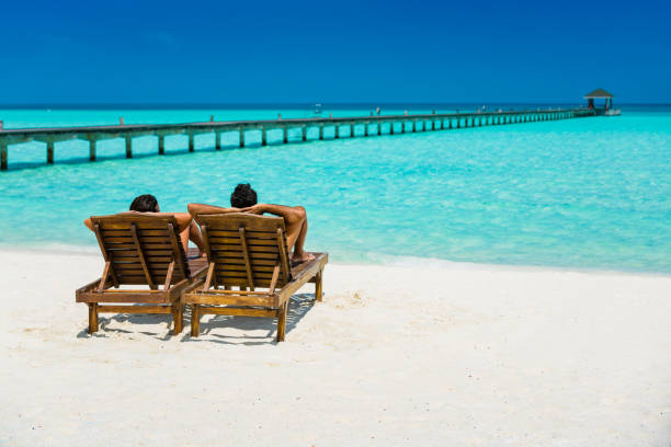 Couple enjoying holidays at Dhiffushi Holiday island, Maldives Couple enjoying holidays sitting on lounge chairs at tropical paradise beach at Dhiffushi Holiday island at South Ari atoll, Maldives. Beautiful turquoise Indian ocean sea with white sand. Luxury travel holidays background. Model and Property released. beach holiday stock pictures, royalty-free photos & images