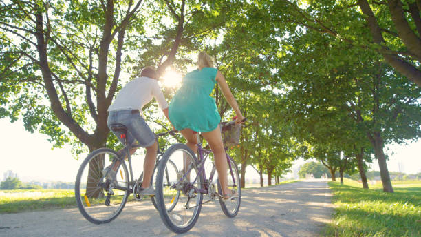 LOW ANGLE: Couple enjoys an active date by riding bikes down a sunlit avenue stock photo