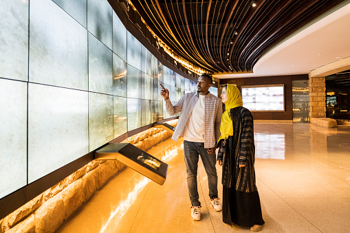 Side view of mid adult tourists standing indoors watching 25 meter video wall at UNESCO World Heritage Site near Riyadh. Property release attached.