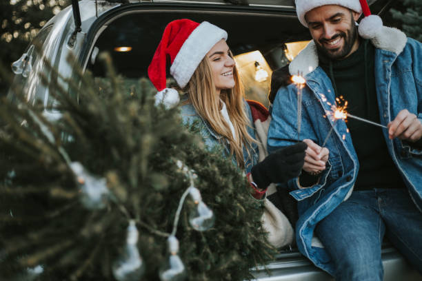 Couple having fun on Christmas tree adventure. Happy couple is wearing Santa Claus hat and sitting in car trunk while enjoying their beautiful moments of Christmas tree adventure. It is a cold and sunny day outside. flora family stock pictures, royalty-free photos & images