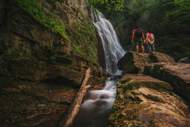 Couple hikers with backpacks enjoying view waterfall in rain forest Couple travelers with backpacks relaxing in greens jungle and enjoying view in waterfall. waterfalls stock pictures, royalty-free photos & images