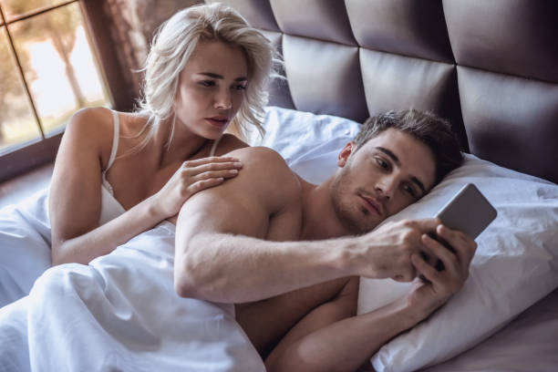 Couple in bed Beautiful young couple is lying in bed, guy is using a smart phone, girl is looking with reproach envy stock pictures, royalty-free photos & images