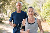 Senior man and woman jogging in park while listening to music. Mature couple running at park together. Retired man and mid woman exercising outdoor.