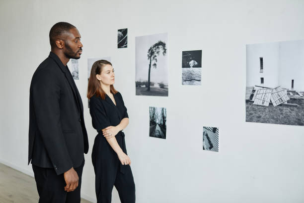 Couple Looking at Photos in Gallery Portrait of elegant mixed-race couple wearing black while looking at photographs in modern art gallery, copy space exhibition photos stock pictures, royalty-free photos & images