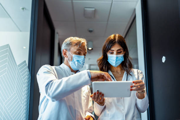 Couple of diverse male doctors walking through corridor looking at tablet. medicine, health and healthcare services. In the Hospital Two Doctors Walking Through the Hospital Hallway, They Talk and Share Tablet Computer. Modern Hospital with Patients and Medical Personnel. physician assistant stock pictures, royalty-free photos & images