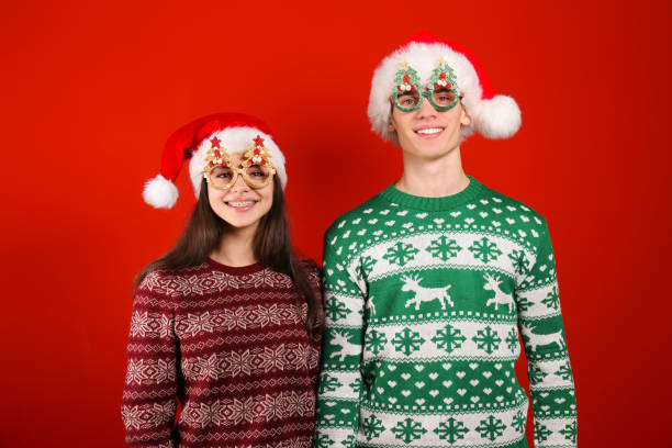 Couple of young people wearing ugly christmas sweaters, posing over colorful isolated background. Studio portrait of young couple, boyfriend & girlfriend wearing santa claus hat & ugly christmas sweater. Holiday outfit w/ snowflake pattern print. Close up, copy space for text, isolated background. ugly girl stock pictures, royalty-free photos & images