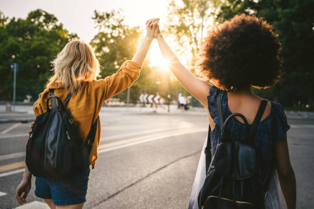 Couple of young women from the back, holding hands with arms raised and they walk in the street at sunset - Two millennials are happy in the city stock photo