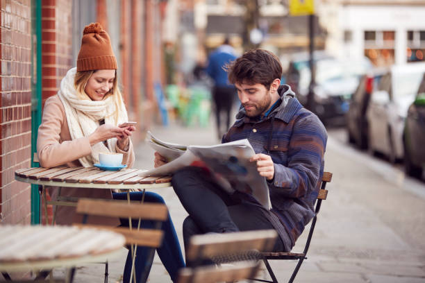 Couple On Date Sitting Outside Coffee Shop On High Street Using Mobile Phone And Reading Newspaper  mid adult couple stock pictures, royalty-free photos & images