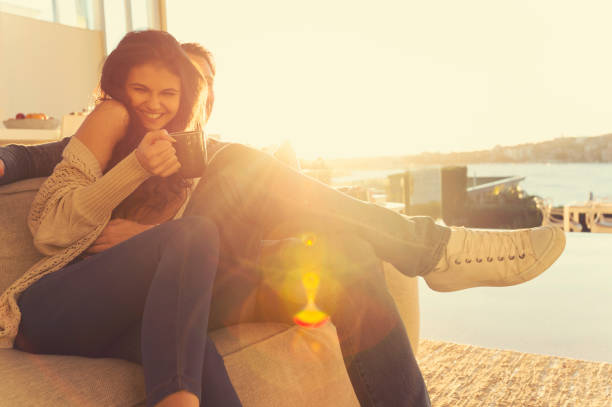 Couple on the sofa at sunrise. Couple on the sofa at sunrise. The woman has a cup of coffee. They are in a luxury house back lit by the sun with lens flare. tickling beautiful women pictures stock pictures, royalty-free photos & images