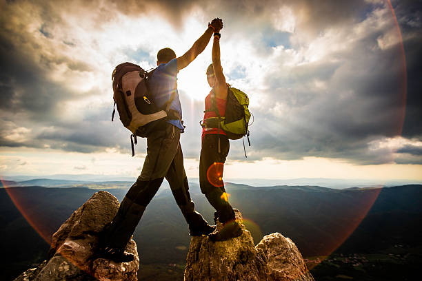 Couple on Top of a Mountain Shaking Raised Hands Couple hiker on top of mountain looking at beautiful sunset landscape, lens flare. extreme sports stock pictures, royalty-free photos & images