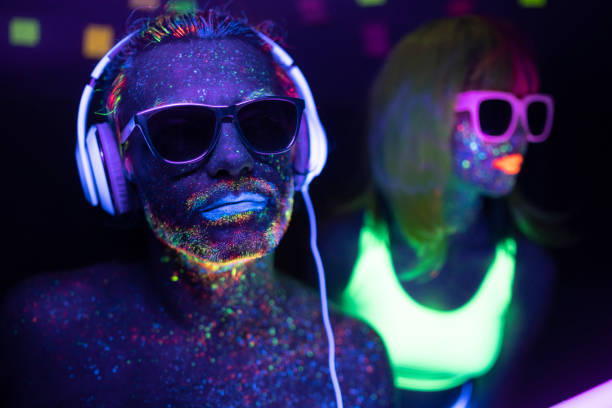 Couple painted in fluorescent UV colors with sunglasses, man with headset Couple with glowing makeup in black light. Young couple with neon makeup powder on face, with sunglasses and headset. paint neon color neon light ultraviolet light stock pictures, royalty-free photos & images