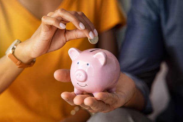 Couple saving money in piggybank Close up of man holding pink piggybank while woman putting coin in it. Indian young couple saving money for their wedding. Close up of woman hand putting euro money in piggy bank to save for the purchase of an house. investment stock pictures, royalty-free photos & images