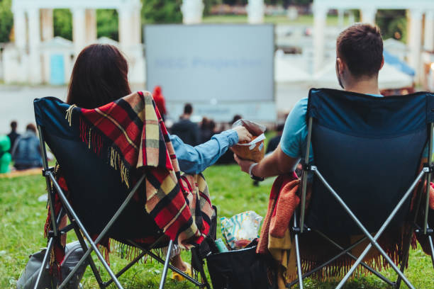 couple sitting in camp-chairs in city park looking movie outdoors at open air cinema couple sitting in camp-chairs in city park looking movie outdoors at open air cinema lifestyle movie theater stock pictures, royalty-free photos & images