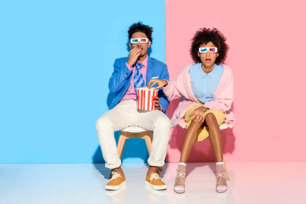 couple sitting on chairs and eating popcorn young african american couple sitting on chairs and eating popcorn against pink and blue wall 3 d glasses stock pictures, royalty-free photos & images