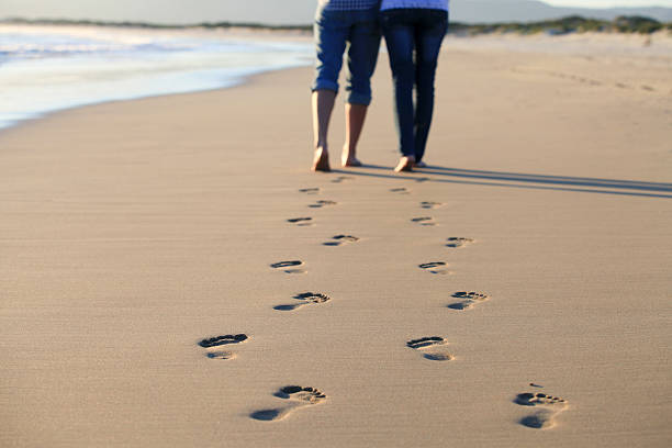 couple walking across beach shoreline leaving footprints - voeten in het zand stockfoto's en -beelden