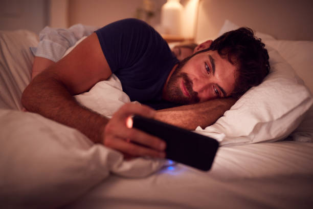 Couple With Man Lying In Bed At Night Looking At Mobile Phone Screen Couple With Man Lying In Bed At Night Looking At Mobile Phone Screen fomo photos stock pictures, royalty-free photos & images