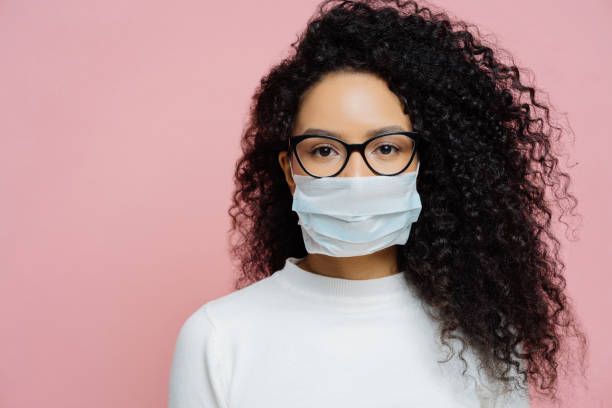 covid-19, infectious virus. close up shot of young woman with curly bushy hair, wears transparent glasses and medical disposable mask, cares about her health, protects in dangerious situation - eyeglasses imagens e fotografias de stock