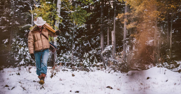 Cowboy carries snowshoes over shoulder while walking through snowy forest stock photo