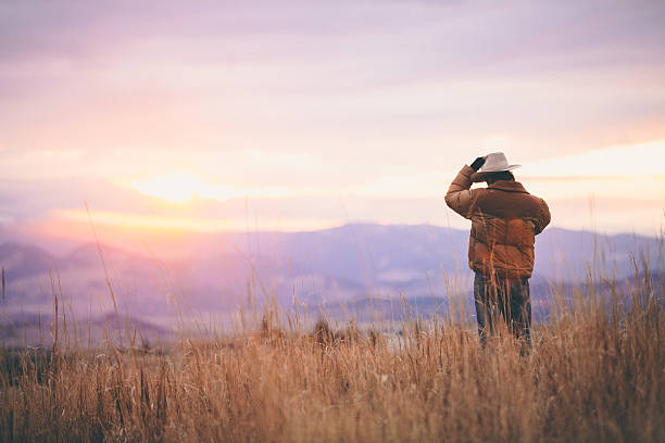 Cowboy talks on phone while watching sunset over mountains stock photo