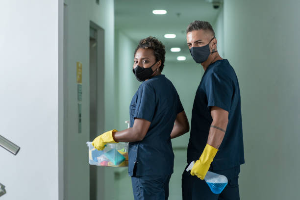 co-workers in charge of cleaning an office walk through their cleaning products while turning and looking towards the camera work team made up of two people, an Afro woman of approximately 45 years of age and a Latin man of 30 years, both with masks for the prevention of covid-19 and dressed in blue toilet uniforms, they are standing in one of the corridor of the offices for those who work with baskets full of toiletries and gloves they stare at the camera that portrays them Office cleaning stock pictures, royalty-free photos & images