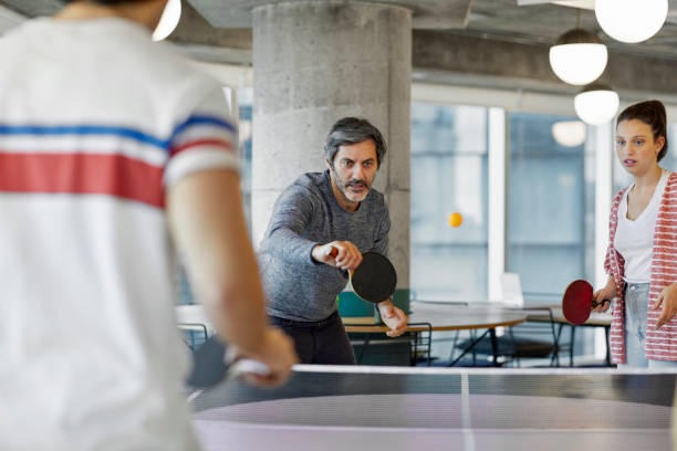 Coworkers playing table tennis in office Mature businessman playing table tennis with colleagues. Male and female coworkers are having fun at workplace. They are in creative office. table tennis stock pictures, royalty-free photos & images
