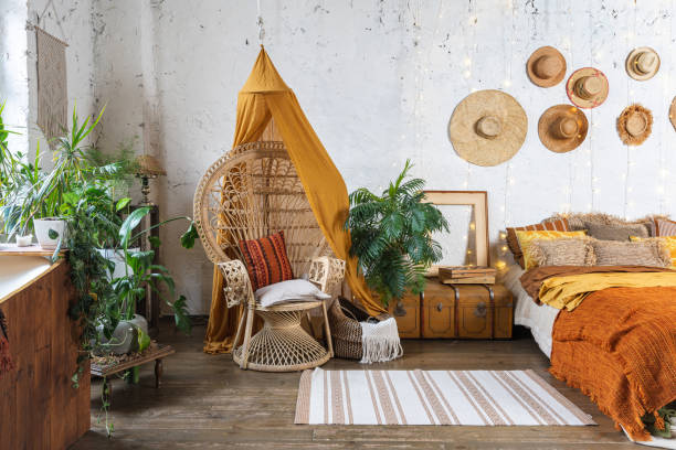 Cozy house with room in boho style interior Elegant and quiet bohemian room with cozy interior, wicker chair, pillows, cushions, green plants in flower pot, bed and rug on wooden floor boho stock pictures, royalty-free photos & images