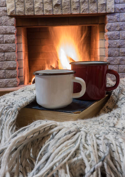 Cozy scene before fireplace with two enameled mugs of tea, a book, and wool scarf. stock photo