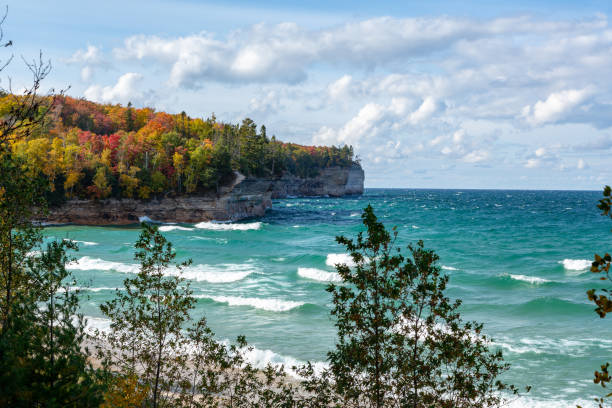 Crashing Waves and Autumn Trees on Lake Superior Shore, Michigan An Upper Peninsula forest creates an autumn background at Chapel Beach in northern Michigan. Lake Superior waves crash on the beach and white puffy clouds dot the blue sky. great lakes stock pictures, royalty-free photos & images