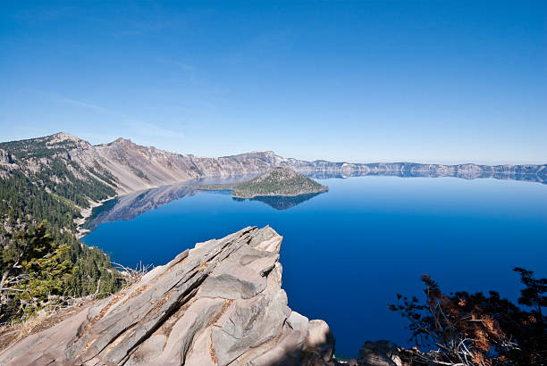 Rock Outcrop Overlooking Crater Lake Crater Lake exists in the blown-out caldera of a once mighty volcano known as Mount Mazama. This view of the lake and Wizard Island was taken from the Rim Trail in Crater Lake National Park, Oregon, USA. jeff goulden crater lake national park stock pictures, royalty-free photos & images