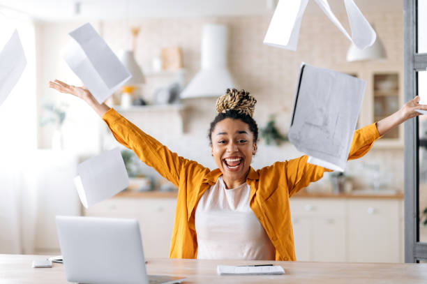 A crazy excited happy young african american woman, business lady, manager or freelancer working remotely at home, enjoying career growth or good deal, gesturing with hands, scattering documents stock photo