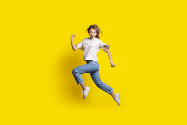 Crazy woman is jumping in a yellow studio wall smiling at camera gesturing a run stock photo