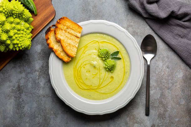 Creamy romanesco broccoli soup with olive oil and toast bread. Vegan food. Fresh raw roman broccoli on background. Directly above. Dark grey table surface. stock photo