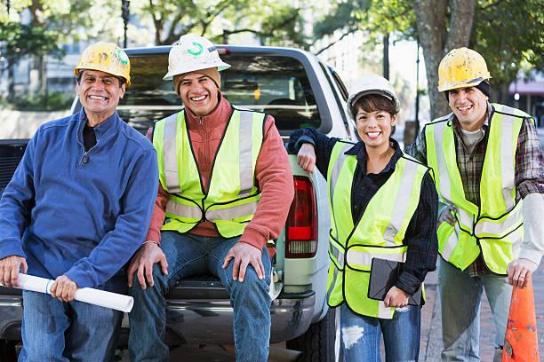 Crew of workers with hardhats and safety vest in city A group of four diverse workers wearing hard hats and yellow safety vests with pickup truck on city street.  They are a crew of construction or utility workers, smiling at the camera.  One of the workers is a woman and two are Hispanic. crew stock pictures, royalty-free photos & images