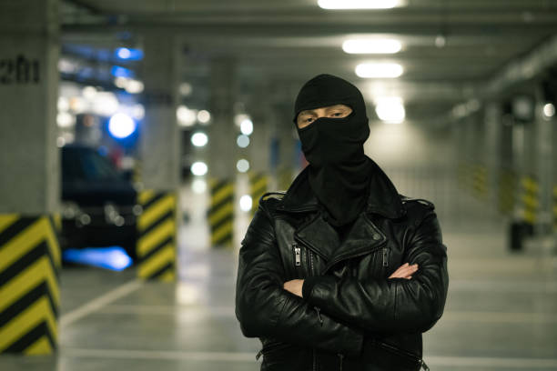 Criminal in black leather jacket and balaclava on head crossing arms by chest Contemporary criminal in black leather jacket and balaclava on head crossing arms by chest with parking area on background ski mask criminal stock pictures, royalty-free photos & images