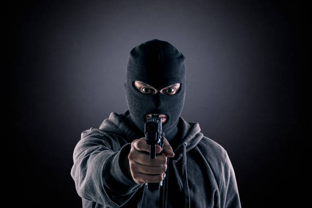 Criminal wearing black balaclava and hoodie with a gun in the dark stock photo