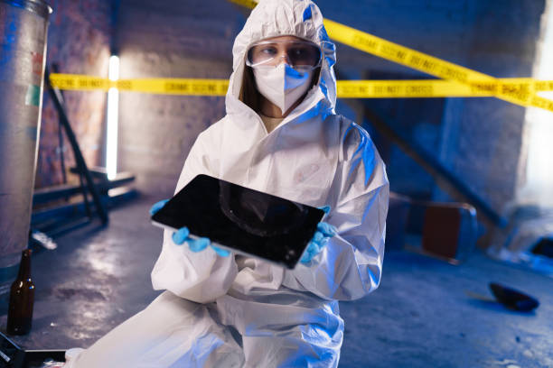 Criminologist in a protective suit against the barricade tape in abandoned warehouse shows a  digital tablet in camera stock photo