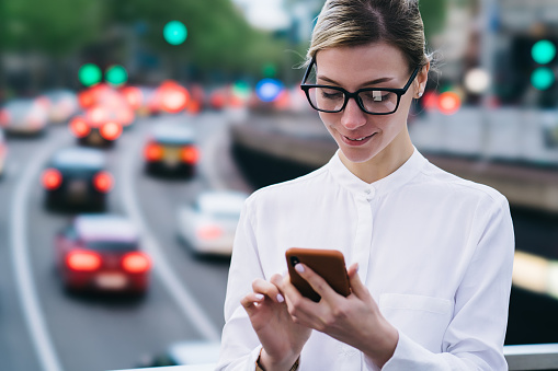 Crop young smiling female in formal outfit and glasses looking at screen while standing on city bridge and messaging on mobile phone in daytime