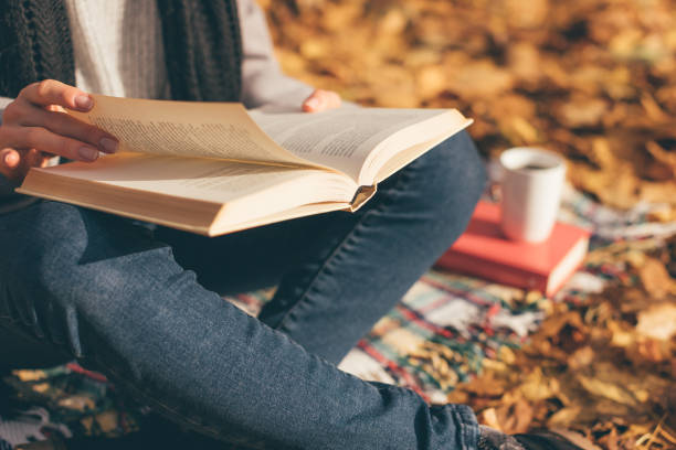 cropped image of young woman sitting on blanket, reading book and drinking coffee or tea in autumn garden - reading imagens e fotografias de stock