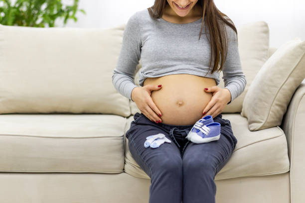 Cropped photo of pregnant woman sitting on the sofa with child clothes. stock photo