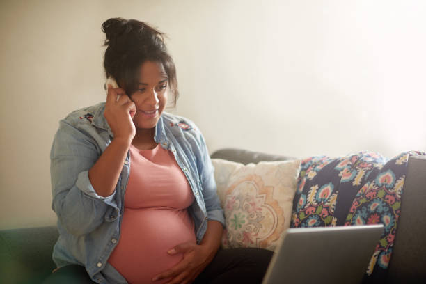 Cropped shot of an attractive young pregnant woman using her cellphone and laptop while sitting in her living room at home stock photo