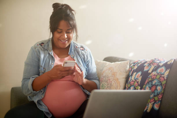 Cropped shot of an attractive young pregnant woman using her cellphone and laptop while sitting in her living room at home stock photo