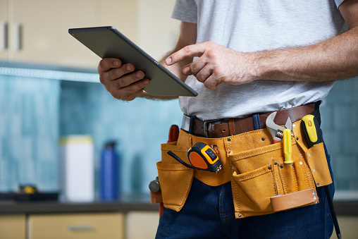 Cropped shot of young repairman wearing a tool belt with various tools using digital tablet while standing indoors. Repair service concept. Horizontal shot